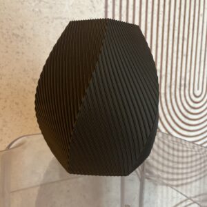 Crna 3D printana vaza na stolici ispred bijele pozadine s geometrijskim uzorkom