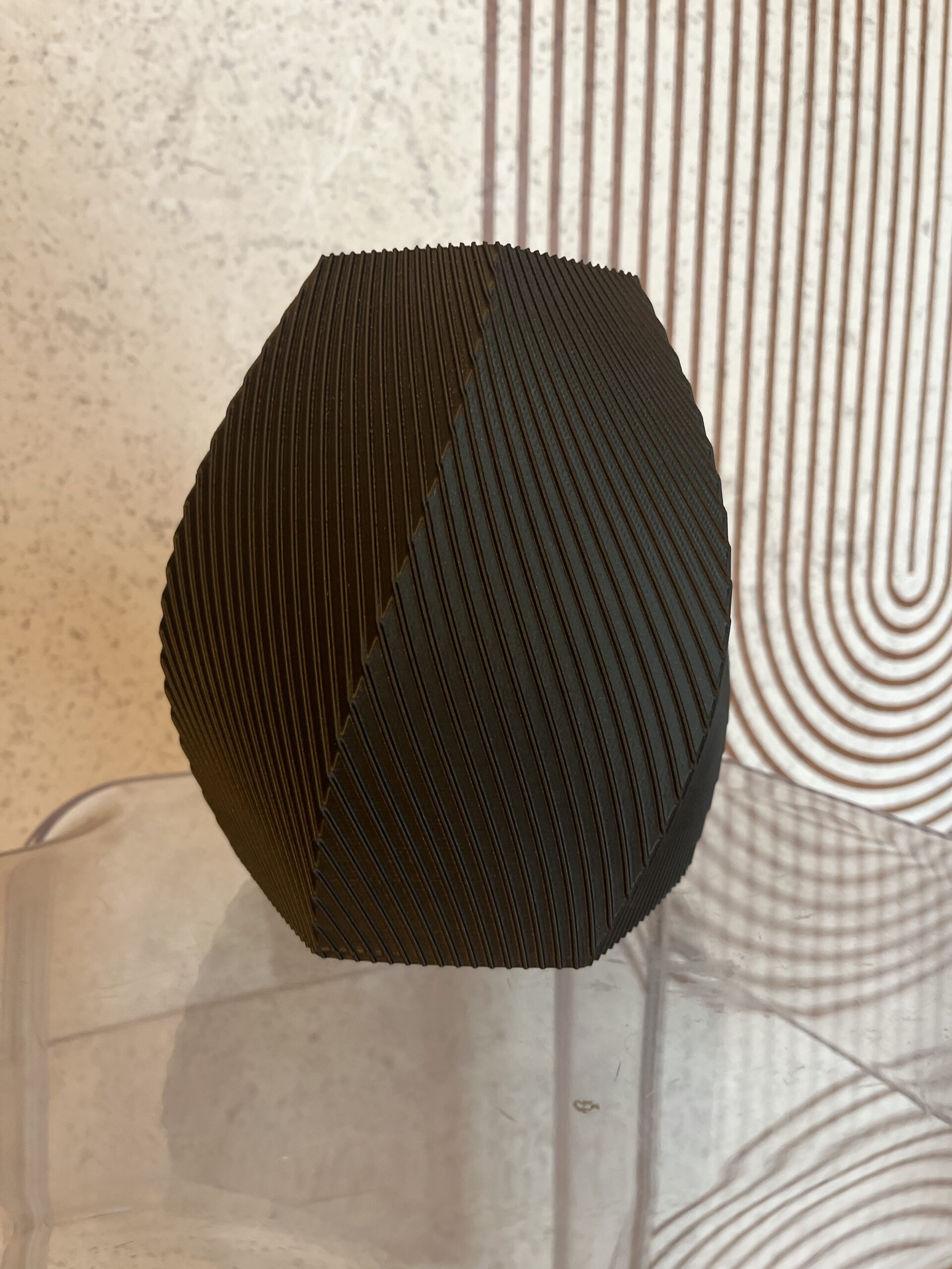 Crna 3D printana vaza na stolici ispred bijele pozadine s geometrijskim uzorkom